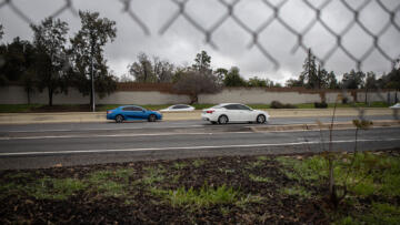 A side-view, as seen through a partially broken wire fence, of cars zooming through a highway on a cloudy day.
